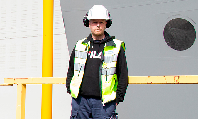 Työnjohtaja Jarkko Kujala seisoo teollisuushallin edessä katsoen kameraan, kädet taskussa, kypärä päässään ja huomioliivi päällä.