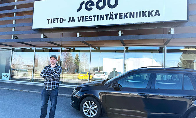 Ohjelmistokehittäjä-opiskelija seisoo ulkona auton vieressä ja rakennuksen edessä, jossa lukee "Sedu, tieto- ja viestintätekniikka".